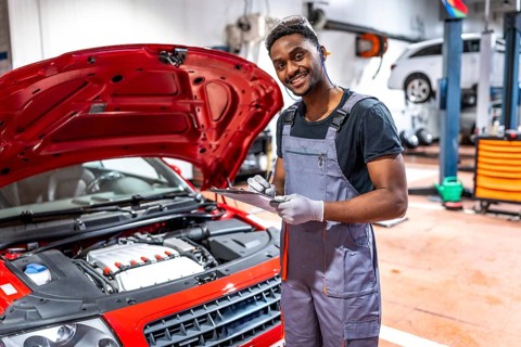Smiling mechanic taking notes on clipboard while inspecting engine of red car with open hood in a