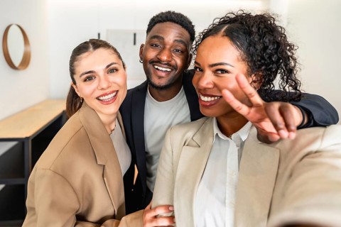 Three cheerful businesspeople taking a selfie, with one making victory sign, celebrating success in