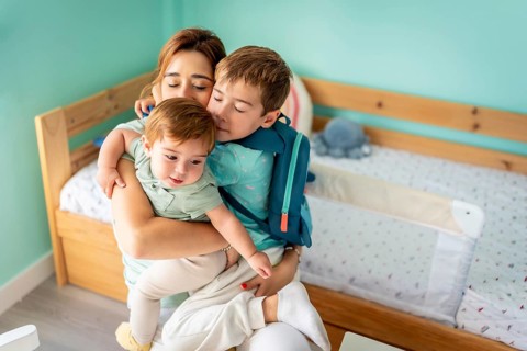 Happy family of three. Mother embracing boy and baby sitting in the bedroom