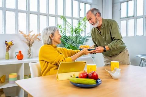 Senior man serving breakfast to share with his wife while she is using digital tablet sitting on
