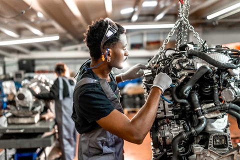 Young mechanic repairing a car engine hanging from a chain hoist, wearing safety goggles and