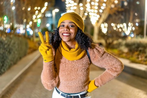 Smiling woman wearing yellow winter clothes showing victory sign while enjoying christmas lights in