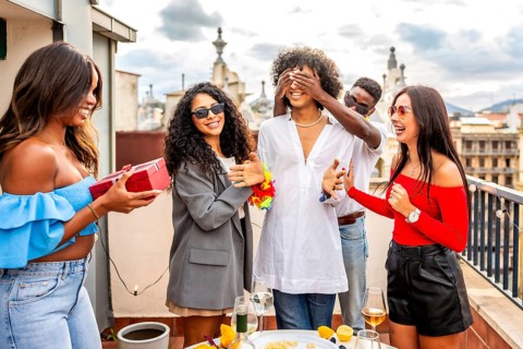 Multiracial group of friends covering eyes of a man giving her a present in a rooftop party