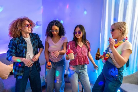 Four friends are singing karaoke and dancing together at a house party, enjoying music, drinks, and