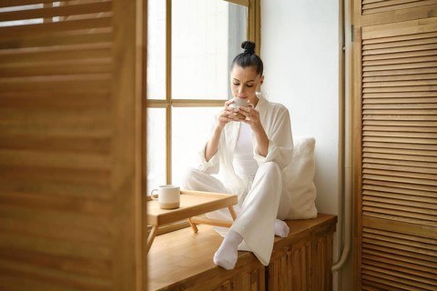 Pretty woman enjoys a peaceful morning, sipping coffee while sitting on windowsill, basking in the