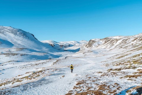 A hiker in a yellow windbreaker on the snow-covered trail to the Reykjadalur hot spring river in