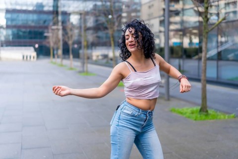 Curly haired hispanic woman dancing in urban city street with modern buildings in the background,
