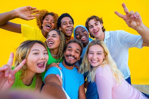 Multi-ethnic friends gesturing peace while taking a selfie next to a yellow wall