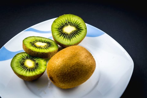 One whole and two cut kiwi on a white plate with a black background