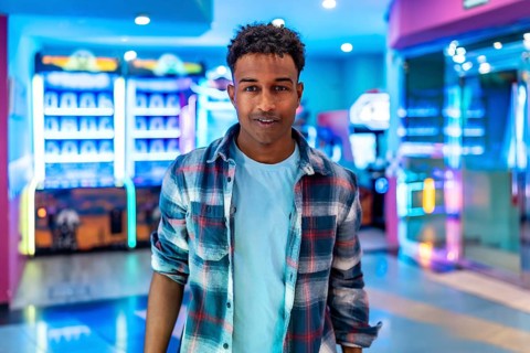 Young man smiles while standing in a lively arcade, surrounded by colorful claw machines and other
