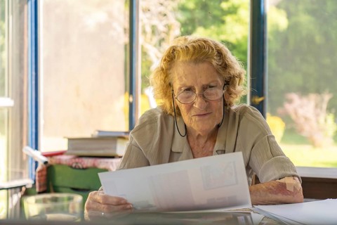 Focused elderly woman with glasses reviewing paperwork, possibly bills or medical records, sitting