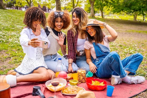Four cheerful young women enjoying a picnic in a park, taking a joyful selfie with a smartphone