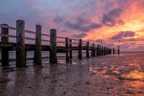 A jetty on the beach at sunset, the sky is coloured orange and purple, the water reflects the