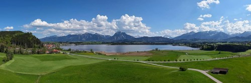 Expansive landscape with a view of Lake Hopfensee and mountains, surrounded by green meadows and a