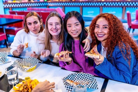 Four diverse young women enjoying a meal together at a restaurant, holding slices of pizza and