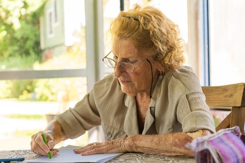 Concentrated elderly woman wearing glasses drawing with green colored pencil on sheet of paper