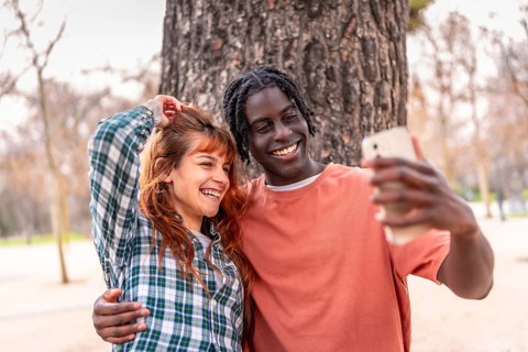 Young multiracial couple embracing and taking a selfie with a smartphone in a park, enjoying a