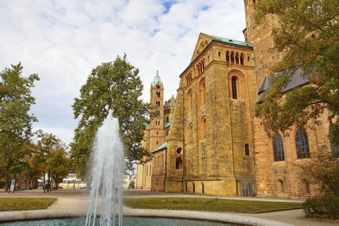 Side view of Speyer Cathedral with towers, trees and a fountain in front of a cloudy sky, Speyer