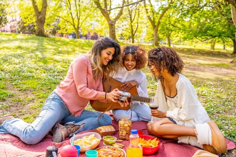 Three friends enjoying a picnic in a park, taking photos with a vintage camera, playing guitar, and