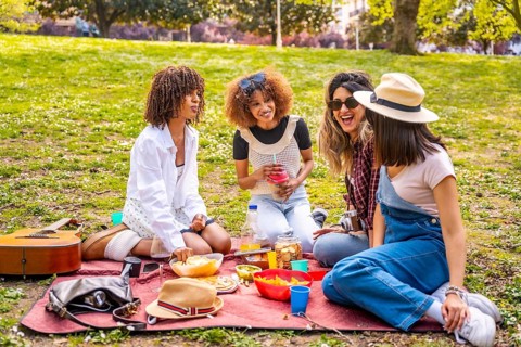 Four diverse female friends enjoying a picnic in a park, sharing food, drinks, and laughter on a