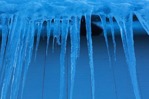 Long icicles and thick heavy ice hanging from roof edge of grey wood plank cladded building in