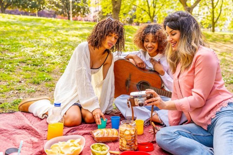 Three female friends enjoying a picnic in a park, taking pictures with a vintage camera, eating