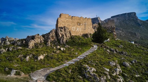 Drone shot, stone ruin on hill with paved path in mountainous landscape under blue sky,