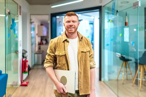 Young professional walking in a modern office corridor, holding a laptop and smiling, showcasing
