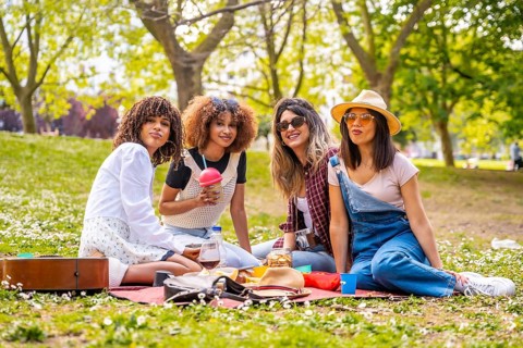 Four diverse female friends enjoying a picnic in a park, sharing food, drinks, and good times on a