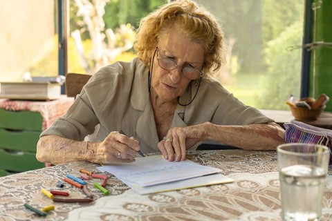 Elderly woman wearing glasses concentrating on drawing with crayons at a table with a glass of