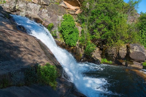 A small waterfall flows over large rocks, surrounded by green plants under a sunny sky, Cascada del