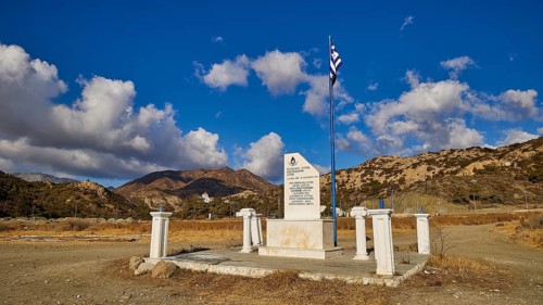 White memorial with Greek flag against a clear blue sky and mountain backdrop, Christopher Litos