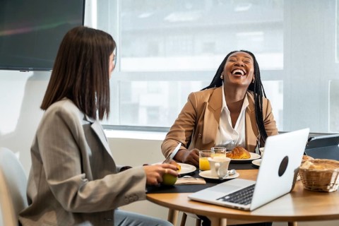 Two businesswomen are enjoying breakfast and laughing together during their business trip, creating