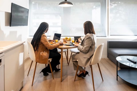 Two businesswomen are enjoying breakfast and working with laptop and tablet in a modern hotel room