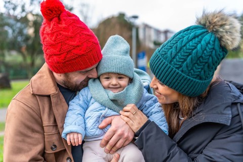 Parents wearing winter hats embracing their smiling baby in a park, enjoying a sunny winter day