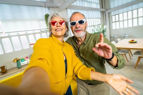 Senior woman with heart shape sunglasses taking selfie with husband while having fun at home