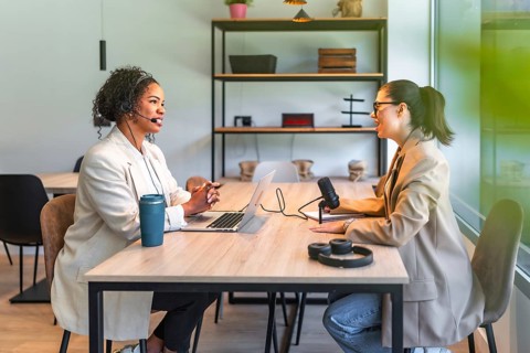 Two female podcasters are recording a new episode in a modern office, using a microphone and laptop