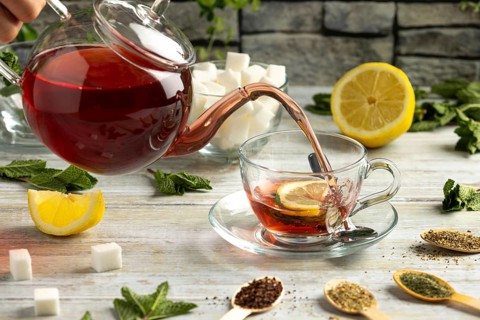 Red tea is poured from a pot into a glass cup with lemon and mint, surrounded by sugar and teas
