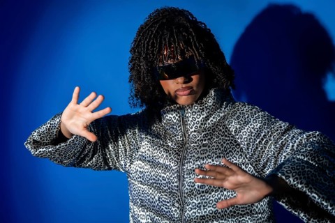 Young woman with curly hair and sunglasses dancing with hands up, wearing a patterned jacket on a