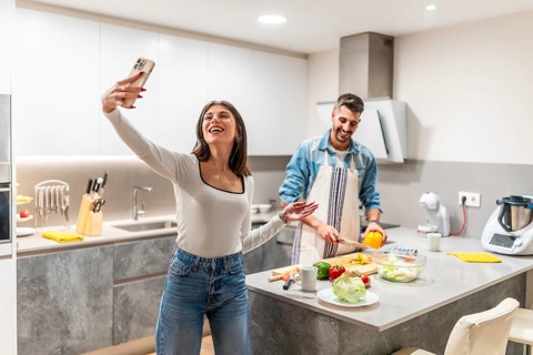 Happy young couple taking selfie with smartphone while preparing healthy food in modern kitchen