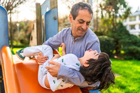 Grandfather catching his grandson sliding down the slide at a playground, having fun together on a