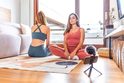 Two women recording an online yoga and meditation class in a cozy living room, embracing wellness