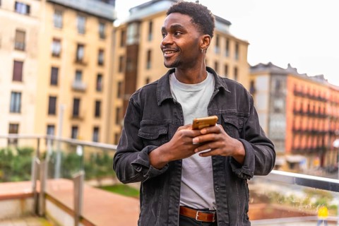 African smiling young man sending a message using smartphone standing int the city