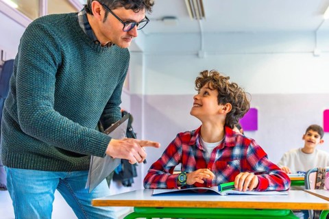 Elementary school teacher engaging with a smiling student while explaining a lesson at their desk