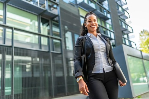 Confident latin modern businesswoman next to block office smiling at camera and carrying laptop and