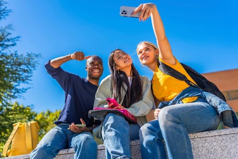 Group of smiling multi-ethnic students taking selfie sitting outside the campus in a sunny day