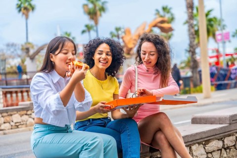 Three female multicultural friends enjoying take away pizza sitting outside the city