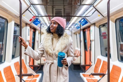 Young black woman wearing winter coat and pink woolen hat holding handrail and reusable coffee cup