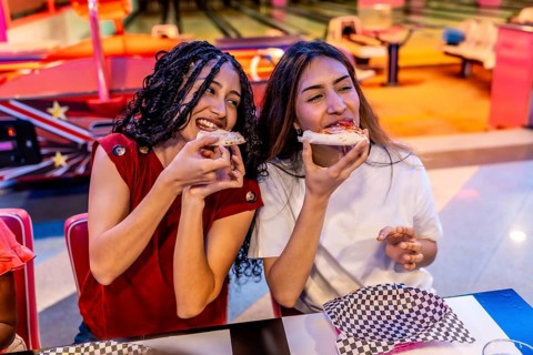 Two joyful young women sharing delicious pizza while enjoying a fun filled day at the bowling