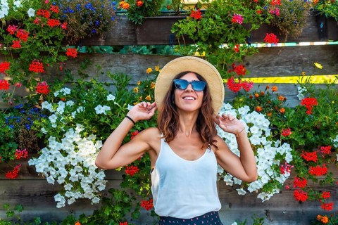 A woman wearing a straw hat and sunglasses is standing in front of a wall of flowers. She is